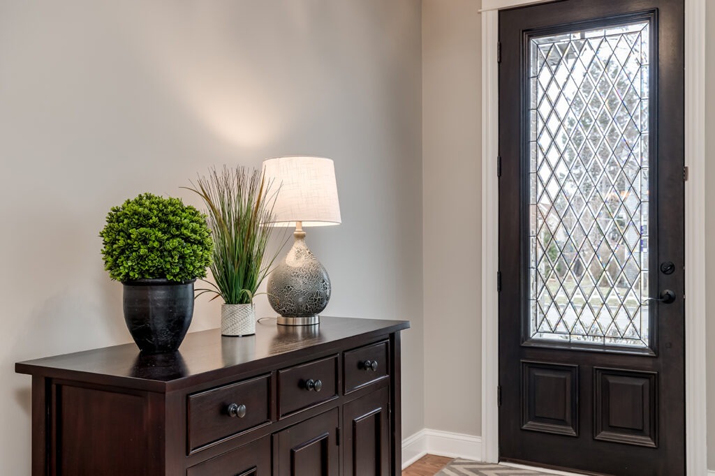Entryway to a large home with plants and a lamp sitting on top of a cabinet with the glass front door behind.