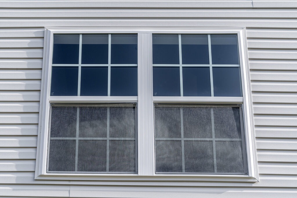 Double hung window with fixed top sash and bottom sash that slides up, sash divided by two white grilles, surrounded by white elegant frame horizontal white vinyl siding on new residence