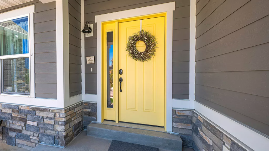 Panorama Yellow front door with wreath and sidelight at the entrance of a house in Utah. Home exterior with gray wood vinyl and stone veneer sidings and a porch with wall lamp near the window.