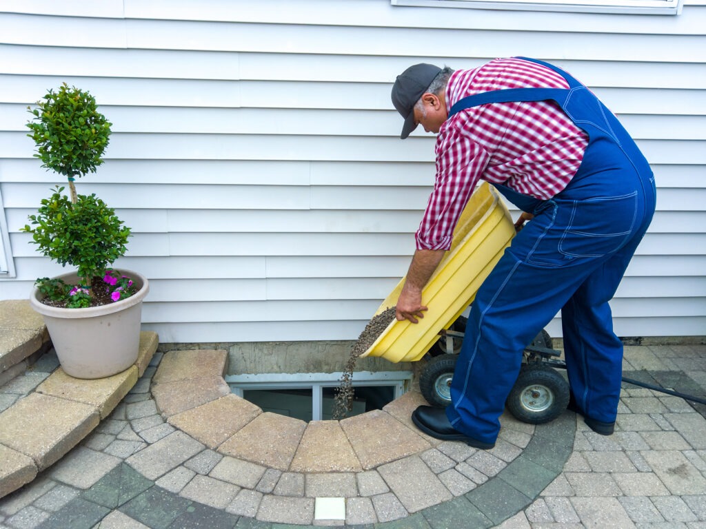 Man doing maintenance on a basement egress window at the side of his house emptying a wheelbarrow full of new pebbles into the cavity surrounded by neat paving