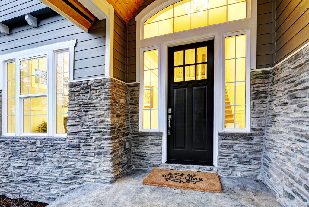 Front covered porch design boasts stone siding which creates immense curb appeal of luxurious home. Welcome mat lead to black front door accented with sidelights framed by white siding.