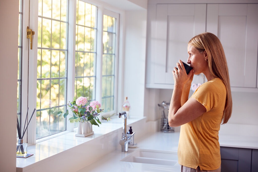 Woman At Home With Hot Drink Standing By Kitchen Window