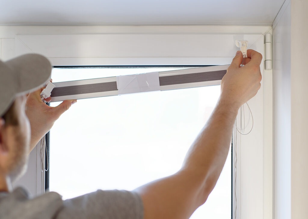 Man installing gray pleated blinds on the window with screwdriver