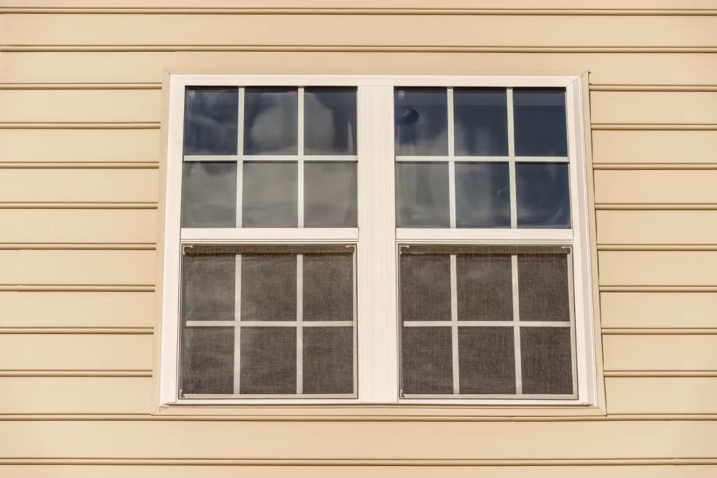 Close up of classic double hung sash window with thin white frame and mutins dividing the window panels, surrounded by desert tan horizontal lap vinyl siding on a new American home in Maryland