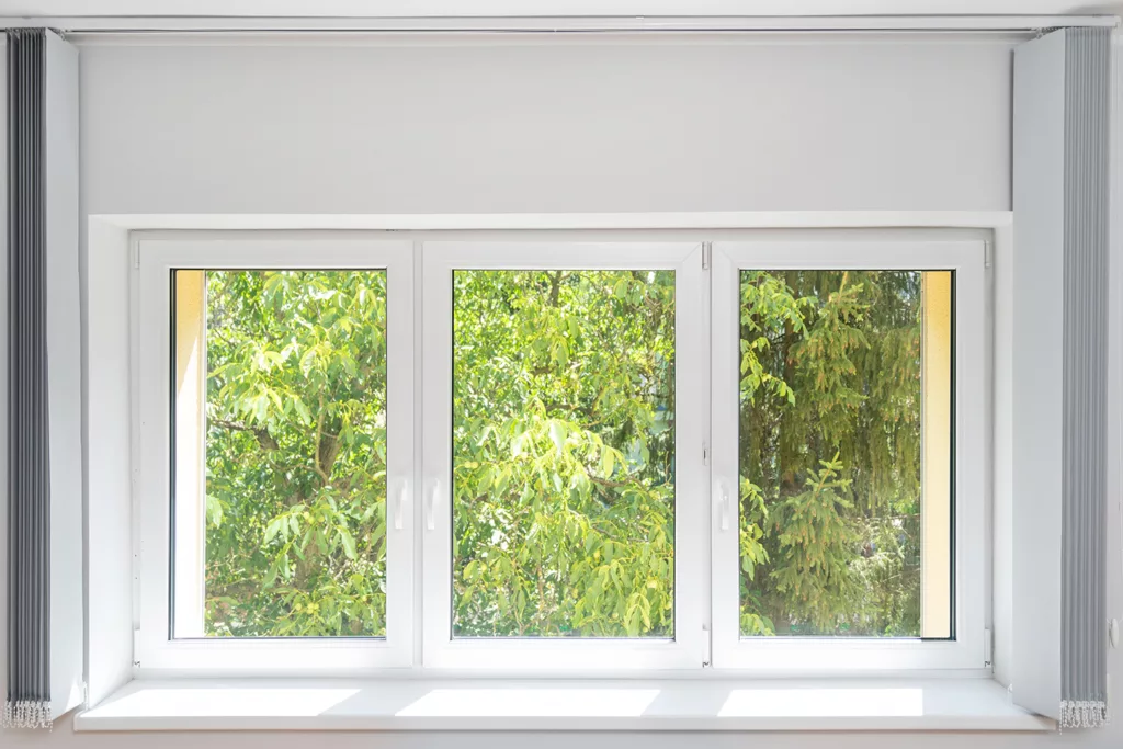 closed plastic pvc window with white frame installed at house, sill and opened vertical fabric jalousie, view on green trees